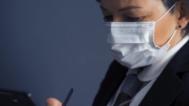 Business woman in a protective mask makes notes in documents holding a tablet in hands wearing black business suit. Business concept. Close-up on head. Prores 422 - Materiał filmowy, wideo