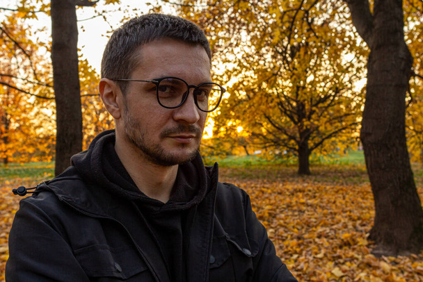 A young short-haired guy wearing glasses and a black jacket stands with his arms crossed in the autumn park. Meditation in the falling park. - Fotó, kép
