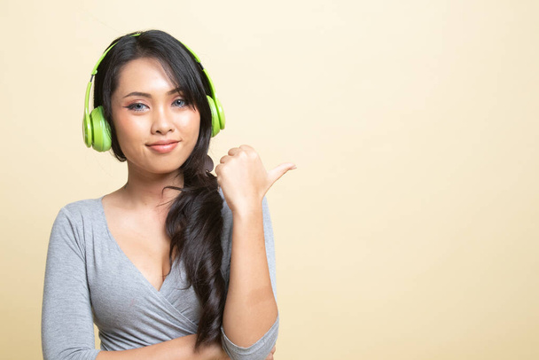 Pretty asian girl listening music with her headphones point to copy space with her thumb   on beige background - Φωτογραφία, εικόνα
