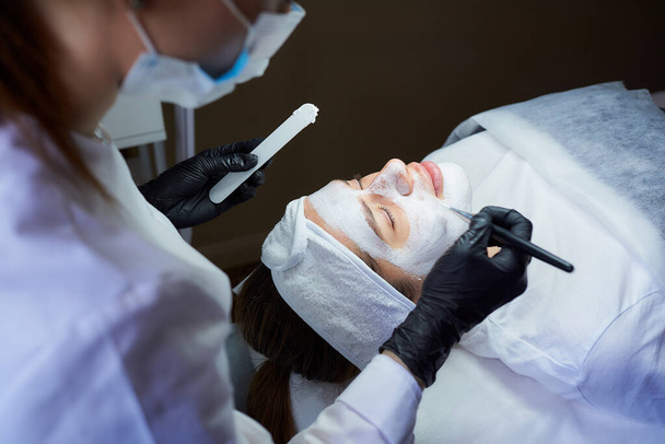Μια πλευρά κοσμητολόγος άποψη που εφαρμόζει ξεφλούδισμα gommage στο πρόσωπο των κοριτσιών. Γιατρός με μάσκα προσώπου και μαύρα αναλώσιμα ιατρικά γάντια κατά τη διάρκεια επέμβασης σε σαλόνι ομορφιάς για καθαρισμό δέρματος - Φωτογραφία, εικόνα