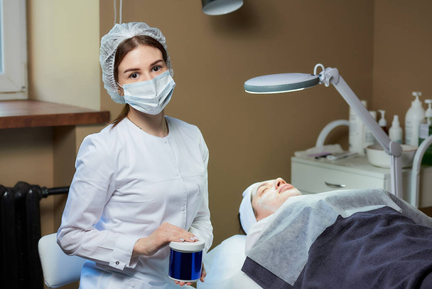 A female cosmetologist in a medical face mask posing with a skincare product near a patient on a full-electrical beauty bed in a cosmetology office. A procedure for skin cleaning in a beauty salon. - Photo, Image