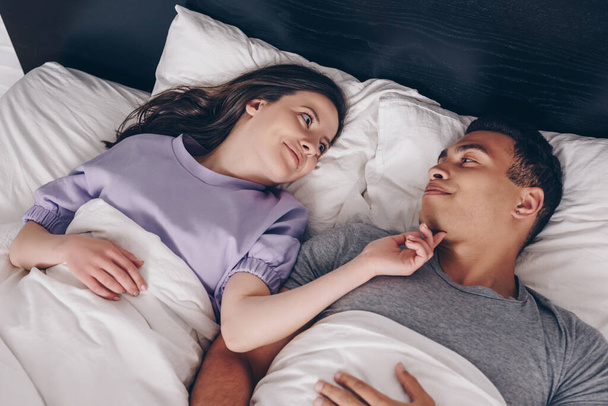 overhead view of cheerful woman touching mixed race boyfriend in bed  - 写真・画像