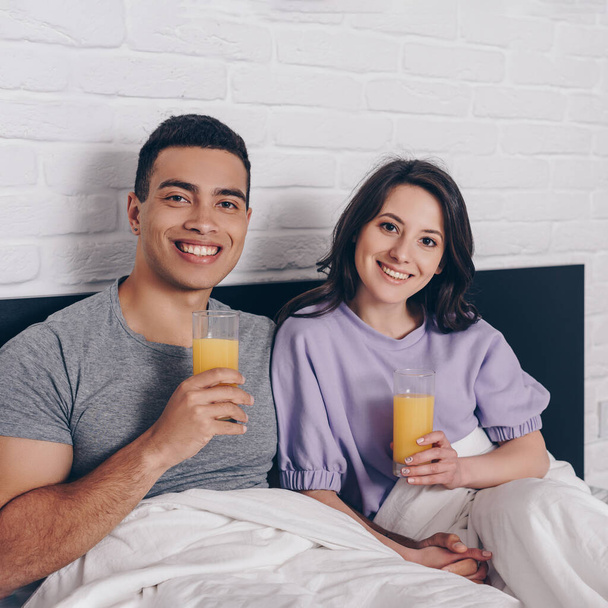 happy mixed race man and cheerful woman holding glasses of orange juice in bed  - Fotó, kép