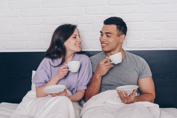 heureux couple interracial tenant des tasses de café et se regardant dans le lit
  - Photo, image
