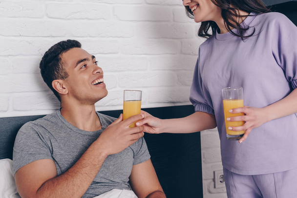 cheerful woman giving glass of orange juice to mixed race boyfriend in bedroom  - Photo, Image