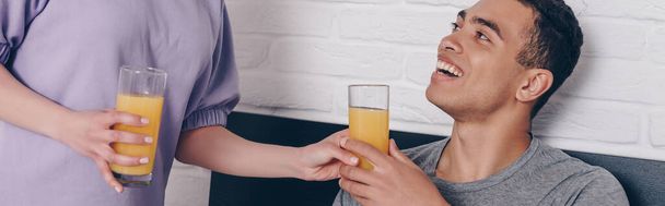 panoramic crop of woman giving glass of orange juice to happy mixed race boyfriend in bedroom  - Fotoğraf, Görsel