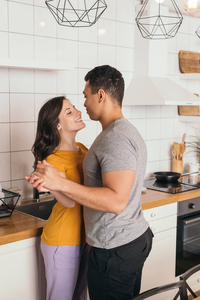 feliz casal interracial de mãos dadas e olhando uns para os outros na cozinha
  - Foto, Imagem