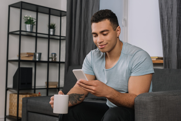 happy mixed race man with tattoo holding cup and using smartphone  - Фото, изображение