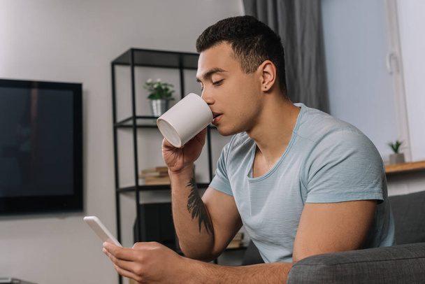 handsome mixed race man with tattoo drinking coffee and using smartphone  - Photo, Image