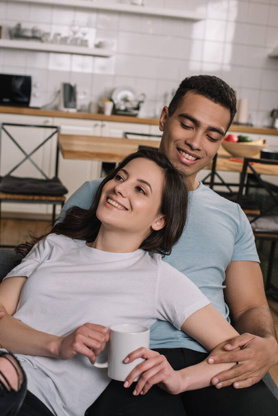 cheerful girl holding cup of coffee near mixed race boyfriend in living room  - Photo, Image