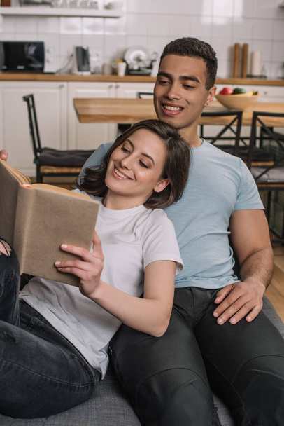 selective focus of happy woman reading book near cheerful mixed race boyfriend - Photo, Image