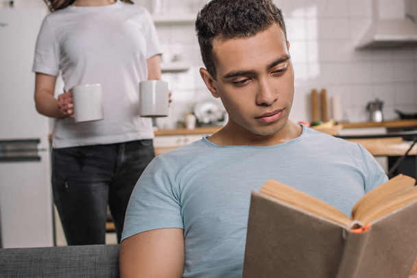 selective focus of mixed race man reading book near girl with cups of coffee - Photo, Image