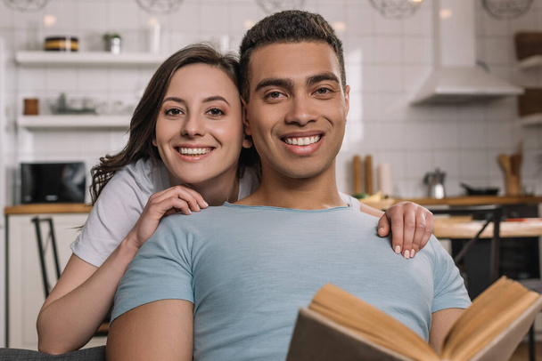 selective focus of handsome mixed race man holding book near beautiful girlfriend  - Фото, изображение