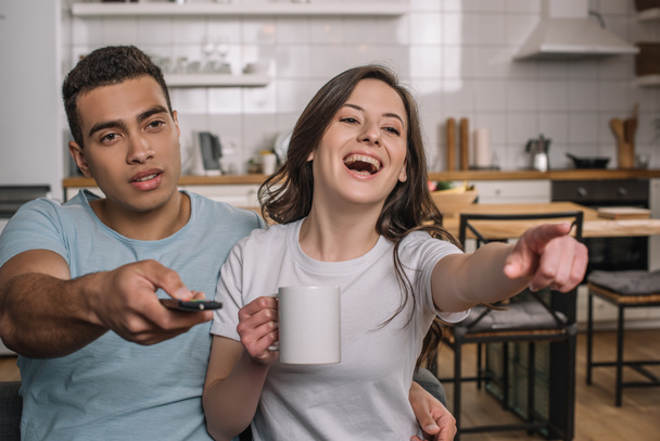 foyer sélectif de l'homme de race mixte tenant télécommande près petite amie heureuse riant et pointant du doigt
  - Photo, image