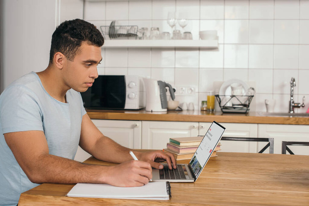 KYIV, UKRAINE - MAY 14, 2020: handsome mixed race man holding pen near notebook and using laptop with airbnb website - Photo, Image