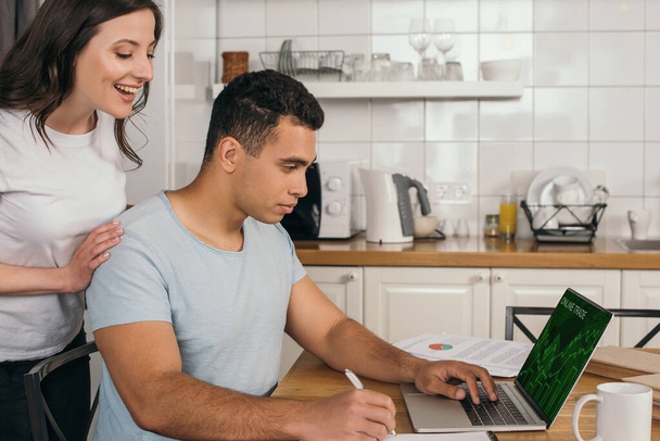 cheerful woman standing near mixed race boyfriend holding pen near laptop with online trade website - Photo, Image
