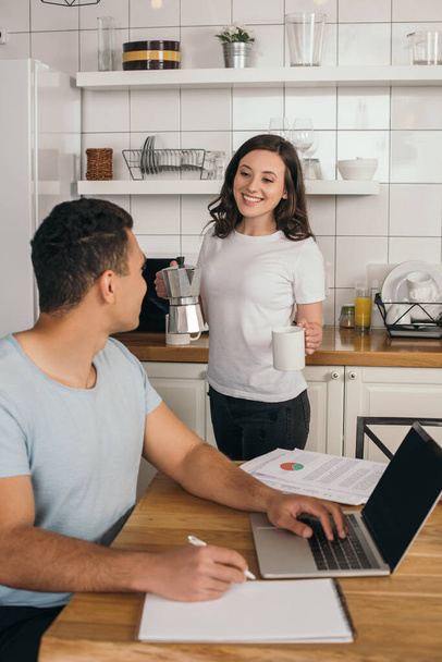 selective focus of cheerful girl holding coffee pot and cup near mixed race man and laptop with blank screen, online study concept  - Photo, Image