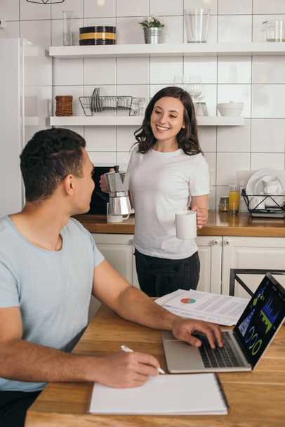 selective focus of cheerful girl holding coffee pot and cup near mixed race man, laptop with charts and graphs - Fotografie, Obrázek