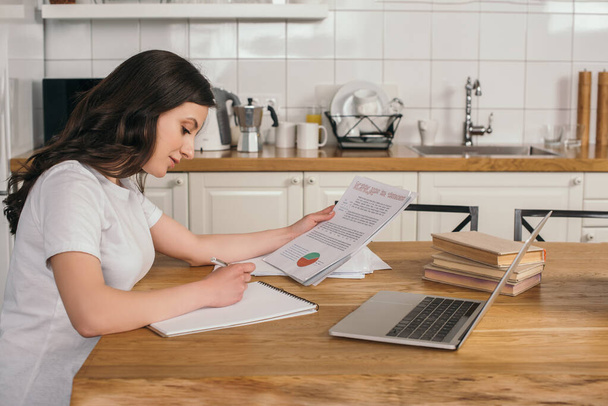 attractive freelancer holding charts and graphs near laptop while writing in notebook  - Fotó, kép