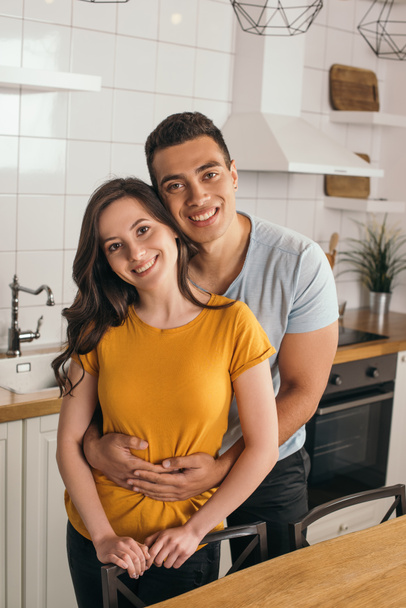 cheerful mixed race man embracing smiling girlfriend in kitchen - Photo, image