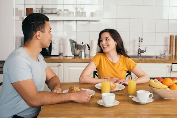 happy woman looking at bi-racial boyfriend near tasty breakfast on table  - 写真・画像