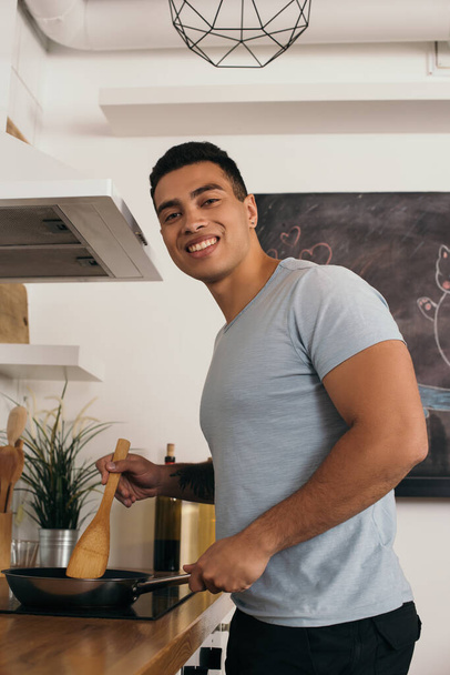 vrolijke bi-raciale man met houten spatel in de buurt van koekenpan  - Foto, afbeelding