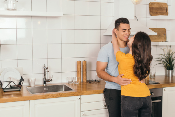 young woman hugging bi-racial boyfriend in kitchen   - Fotografie, Obrázek
