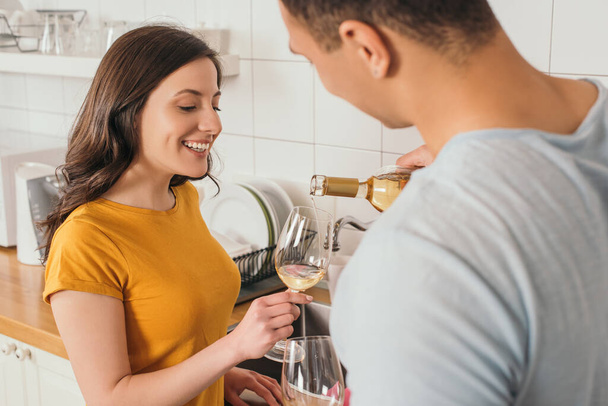 selective focus of bi-racial man holding bottle of wine near glasses and smiling girlfriend  - 写真・画像