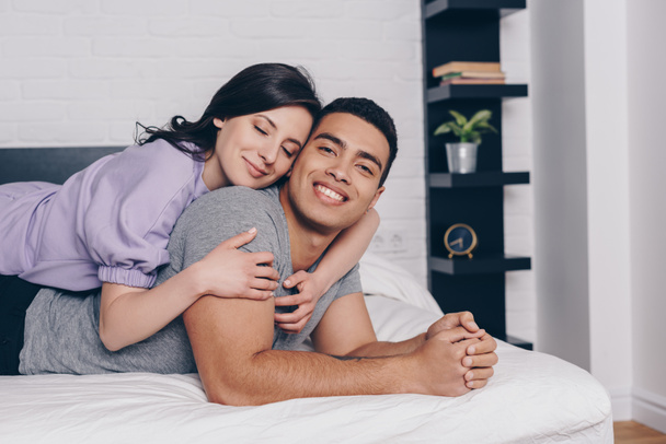 cheerful woman with closed eyes hugging handsome bi-racial man while lying on bed  - Foto, Imagen