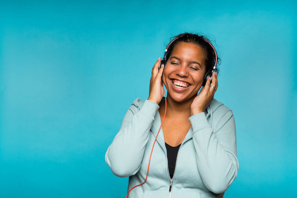 Junge attraktive gemischte Rasse Frau genießt Musik hören mit Kopfhörern blauen Hintergrund - Foto, Bild