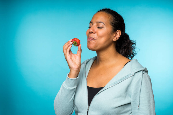 Hermosa joven mujer de raza mixta en ropa casual comiendo y disfrutando de una fresa fresca con un fondo azul
 - Foto, imagen