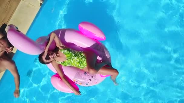 Aerial. Girl chilling on inflatable pink flamingo float in swimming pool. Man relaxing, tanning at poolside. Happy couple chill with floating toys in luxury resort. View from above. Woman in bikini. - Video