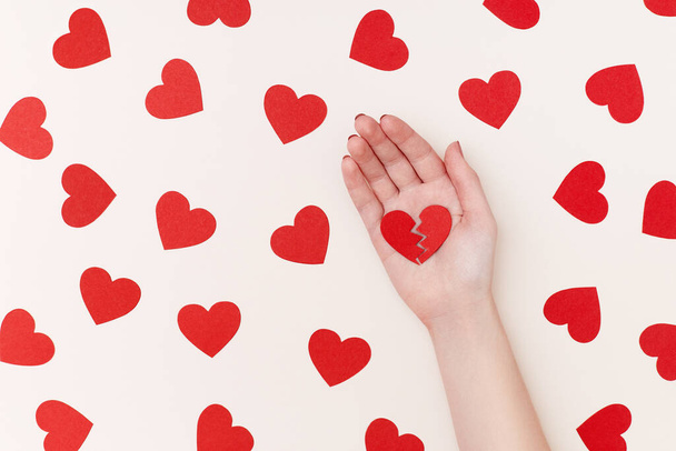 Pattern of red paper hearts surrounding womans hand holding two parts of broken heart on the biege background - Photo, image