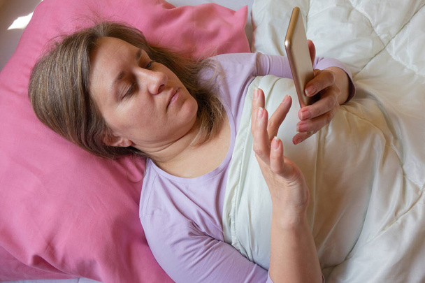 Жінка використовує мобільний телефон у ліжку. Близько 40 років жінка лежить на подушці і смс на смартфоні. Соціальні мережі або залежність від Інтернету
 - Фото, зображення
