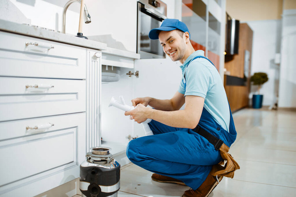 Lächelnder Installateur in Uniform hält Abflussrohr in der Küche. Handarbeiterin mit Werkzeugtasche repariert Waschbecken, Sanitärservice zu Hause - Foto, Bild