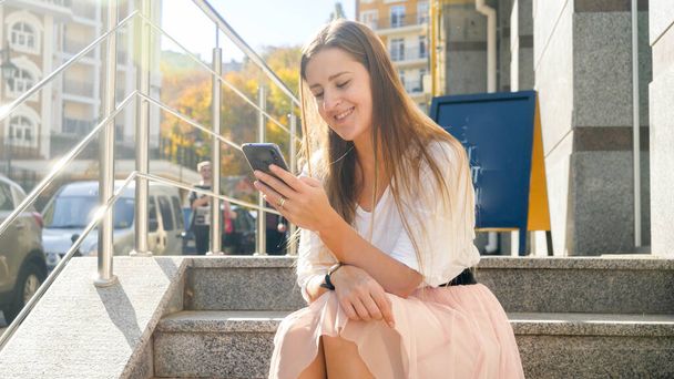 Toned photo of happy smiling girl sitting on stairs on street and browsing internet on smartphone - Фото, изображение