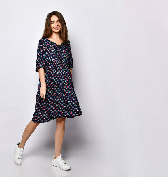 Brunette model in black dress with floral print, white sneakers. Smiling, touching her face, posing isolated on white. Full length - Fotografie, Obrázek