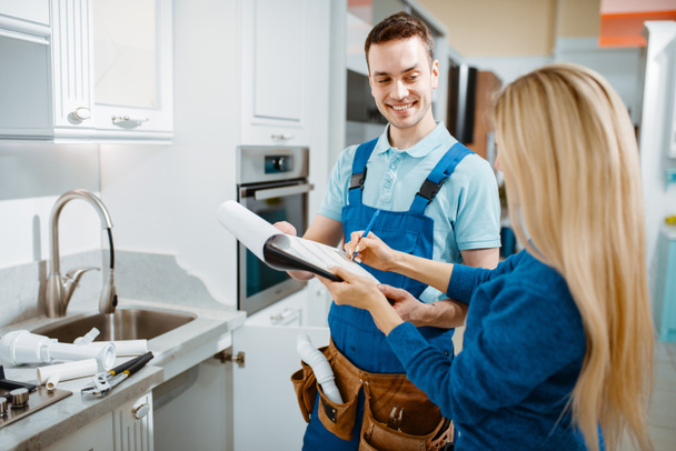 Male plumber in uniform and female customer in the kitchen. Handyman with toolbag repair sink, sanitary equipment service at home - Photo, Image