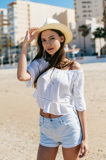 girl in a stylized country costume cowboy stands on the beach sand against the background of high-rise buildings holding a hat with her hand and looking carefully at the camera - Photo, Image