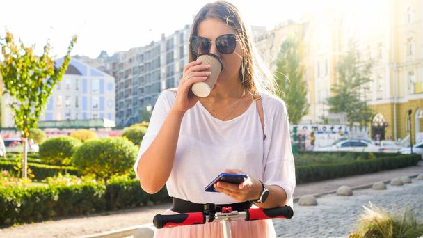 Portrait of beautiful smiling girl in sunglasses drinking coffee and riding on electric scooter on city street - Фото, изображение