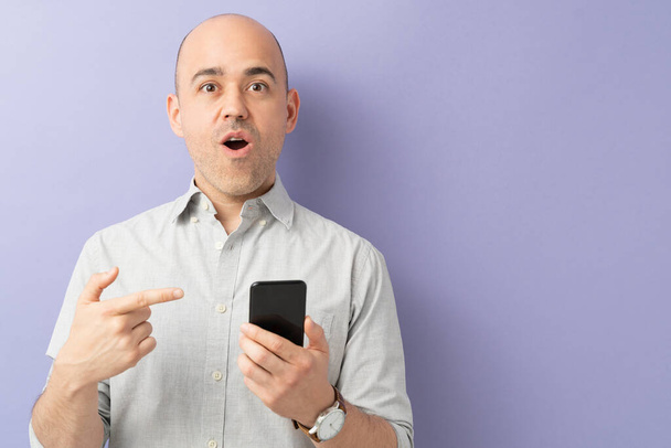 Мужчина смотрит на свой смартфон и выглядит шокированным и удивленным с открытым ртом в студии
 - Фото, изображение