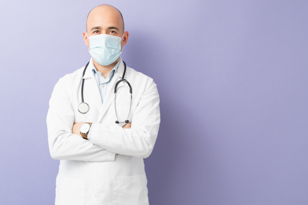 Mężczyzna lekarz w masce chirurgicznej i płaszczu laboratoryjnym z ramionami skrzyżowane w studio z fioletowym tle - Zdjęcie, obraz