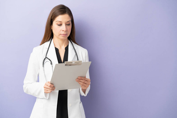 Γυναίκα γιατρός γύρω στα 40 φοράει εργαστηριακή ποδιά και διαβάζει ιατρικό ιστορικό σε στούντιο. - Φωτογραφία, εικόνα