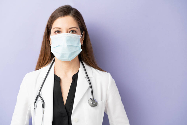 Портрет женщины-врача в маске для лица и халате на студии с фиолетовым фоном
 - Фото, изображение