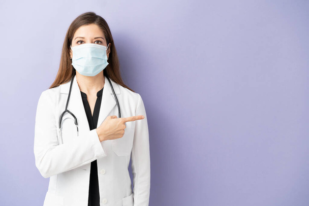 Σοβαρή γυναίκα γιατρός που φοράει μάσκα προσώπου και εργαστηριακή ποδιά που δείχνει προς το χώρο αντιγραφής σε ένα στούντιο - Φωτογραφία, εικόνα