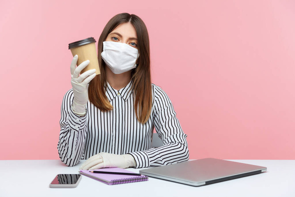 Woman office employee holding disposable cup of coffee, sitting at workplace, feeling safe healthy with hygienic face mask and protective gloves, working in self-isolation, coronavirus quarantine - Фото, изображение