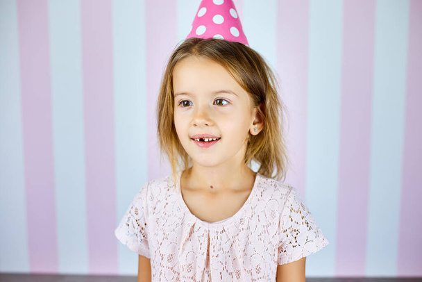 Petite belle fille portant une casquette rose anniversaire sur fond rayé blanc rose souriant avec un visage heureux. Joyeux anniversaire. - Photo, image