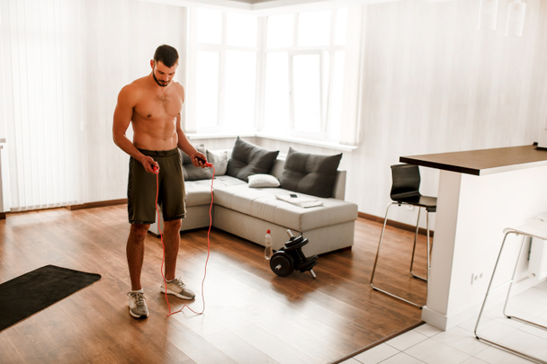 Athletischer Aufbau Ein hübscher Sportler mit nacktem Oberkörper trainiert zu Hause mit einem Springseil. Sportler ohne Hemd treibt Sport im Wohnzimmer - Foto, Bild