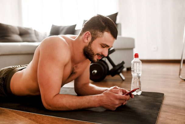 Обычный молодой человек занимается спортом дома. Парень без футболки отдыхает на полу и пользуется смартфоном. Первокурсник в тренировке расслабляющий после тренировки. Один в квартире
 - Фото, изображение