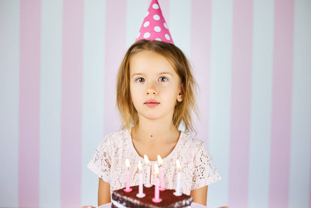 Dziewczynka w różowej czapce urodzinowej z czekoladowym tortem ze świecami. Dziecko świętuje swoje urodziny. Wszystkiego najlepszego. - Zdjęcie, obraz